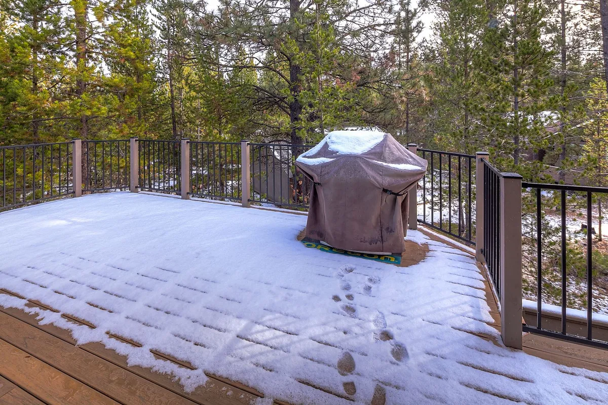 An exterior shot of a deck surrounded by trees, with a grill covered by a protective cover and light snow on the deck boards. The tranquil setting suggests both seasonal versatility and a place to gather outdoors for meals or relaxation while enjoying the natural environment.