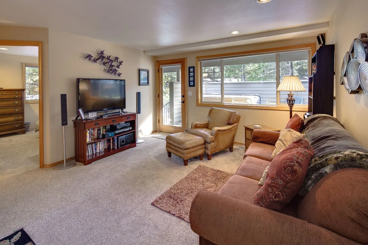 A comfortable family room setting with a sofa adorned with decorative pillows, a television, a reading chair, and a large window letting in natural light. The setup is ideal for relaxation, movie marathons, or quiet downtime.