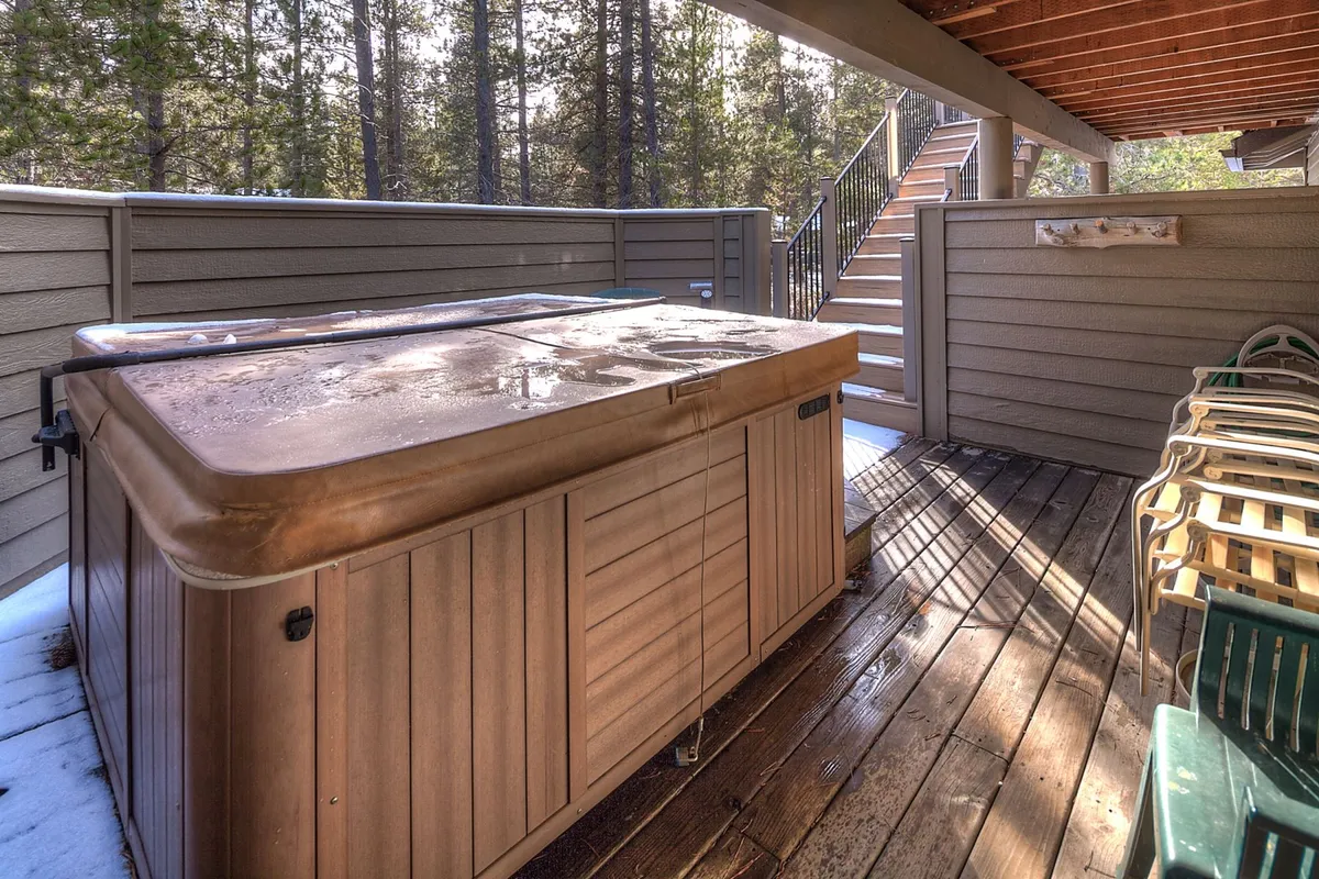 A tranquil lower deck scene featuring a bubbling hot tub encased in a wooden privacy wall. The staircase not only links the two deck spaces but hints at the home’s multi-level outdoor experience. The ambiance is peaceful, inviting guests to soak and unwind while enjoying serene nature views.