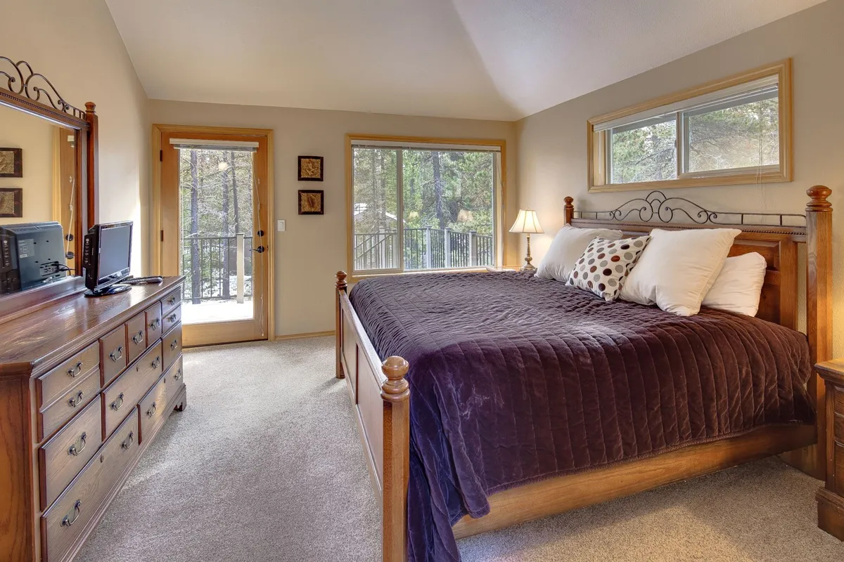A spacious, sunlit bedroom featuring a wooden king-sized bed with plush bedding. Large windows allow natural light to stream in, and a dresser with a mirror offers both function and style. The space exudes comfort and tranquility, ideal for restful nights.