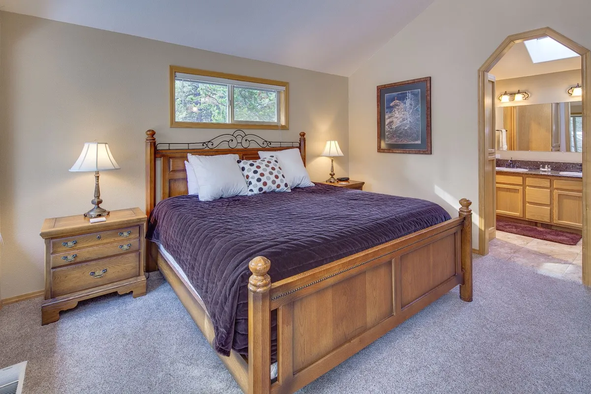 A calm and restful bedroom with a comfortable bed, nightstands with lamps, and artwork on the walls. The bathroom entrance is visible, suggesting convenience and thoughtful design. Sunlight filters gently into the space, enhancing its tranquil feel.