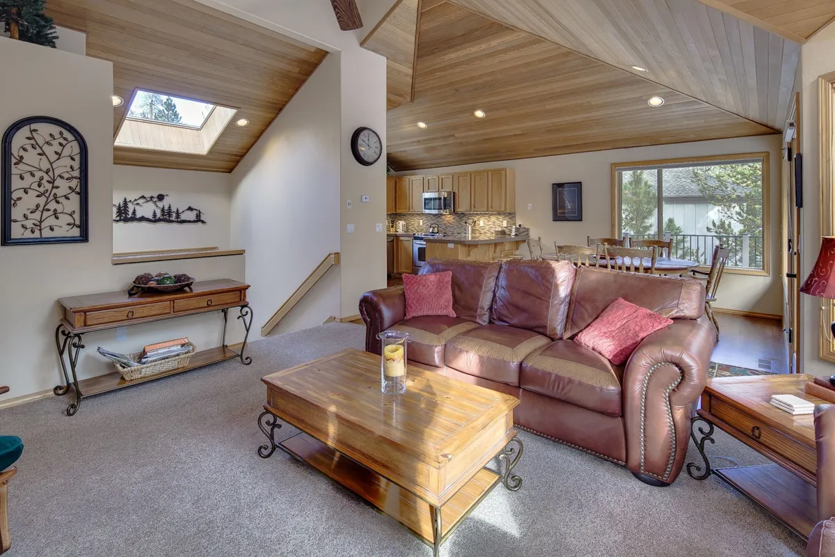 A cozy seating area with a brown leather sofa, wooden coffee table, and a skylight providing abundant daylight. The large window hints at the wooded setting beyond, tying interior leisure to the outdoors.